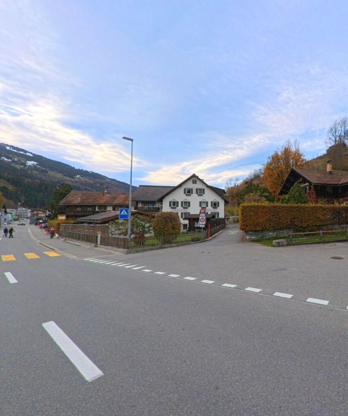 An einer übersichtlichen Kreuzung mit Fussgängerstreifen stehen Wohnhäuser im Dorfbereich von Küblis (7240, Kanton Graubünden) vor einer ruhigen Hanglandschaft.