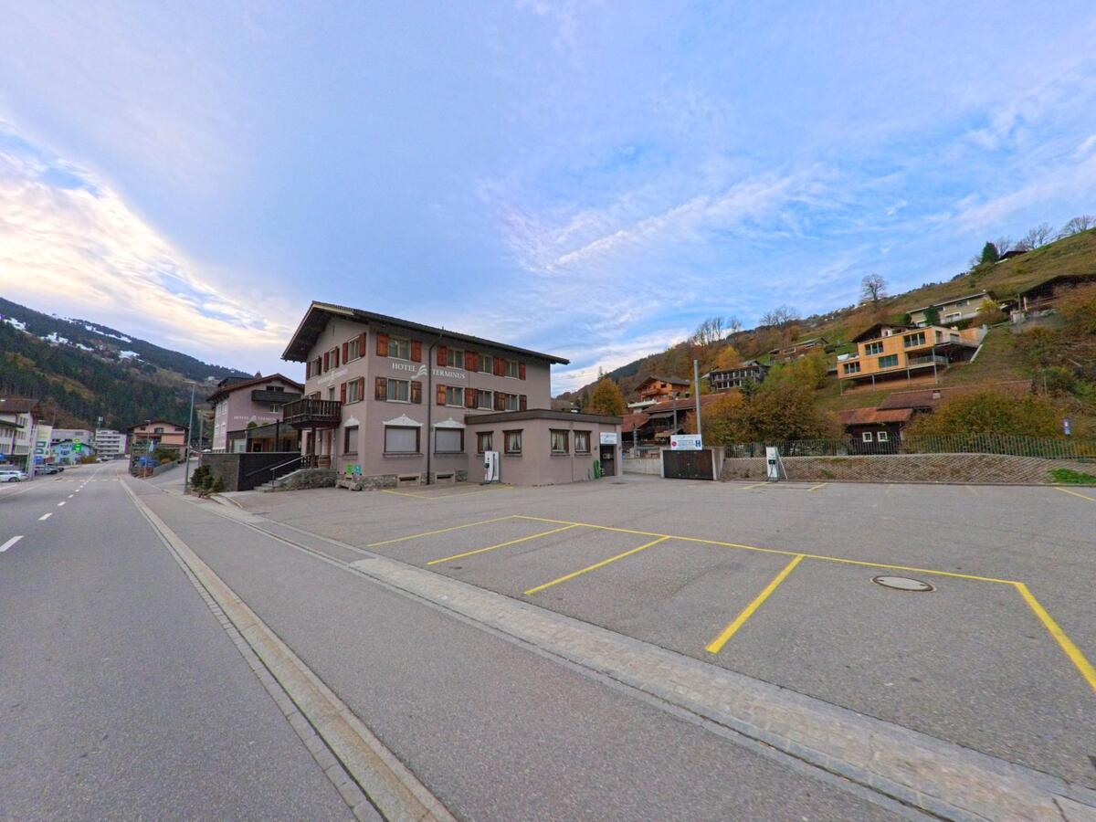 Neben einer breiten Strasse steht ein markantes Gebäude mit Parkplatz im Siedlungsgebiet von Küblis (7240, Kanton Graubünden), eingerahmt von Hanglagen und herbstlicher Umgebung.