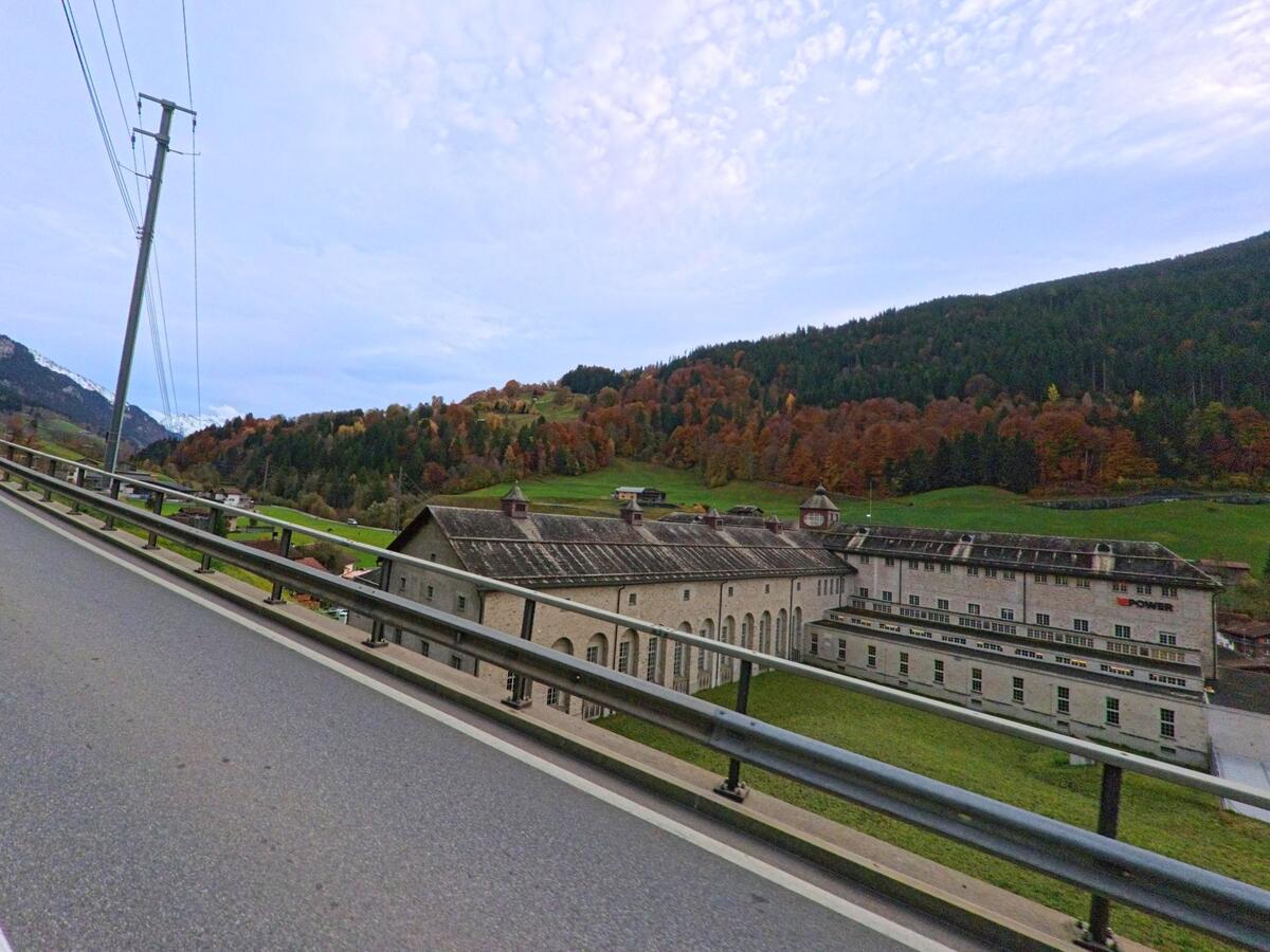 Von einer erhöhten Strasse aus zeigt sich ein langgestrecktes Industriegebäude am Talrand von Küblis (7240, Kanton Graubünden), eingebettet in bewaldete Hänge mit Herbstfärbung.