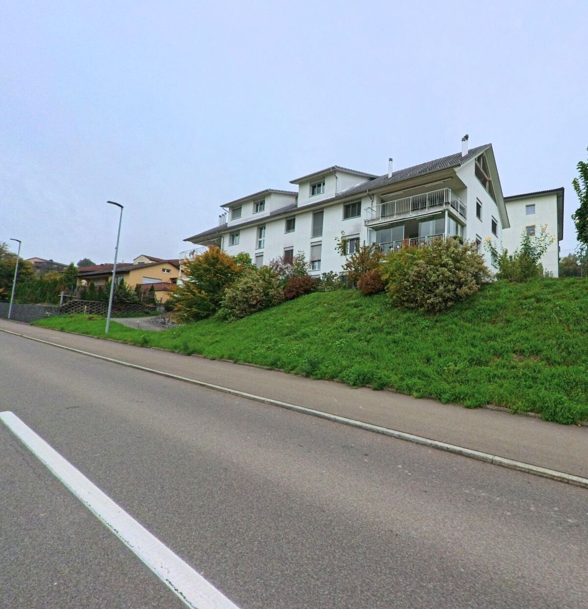 Ein weisses Mehrfamilienhaus in Künten (5444, Kanton Aargau) steht auf einer kleinen Anhöhe über der Strasse, umgeben von Sträuchern und Rasen.