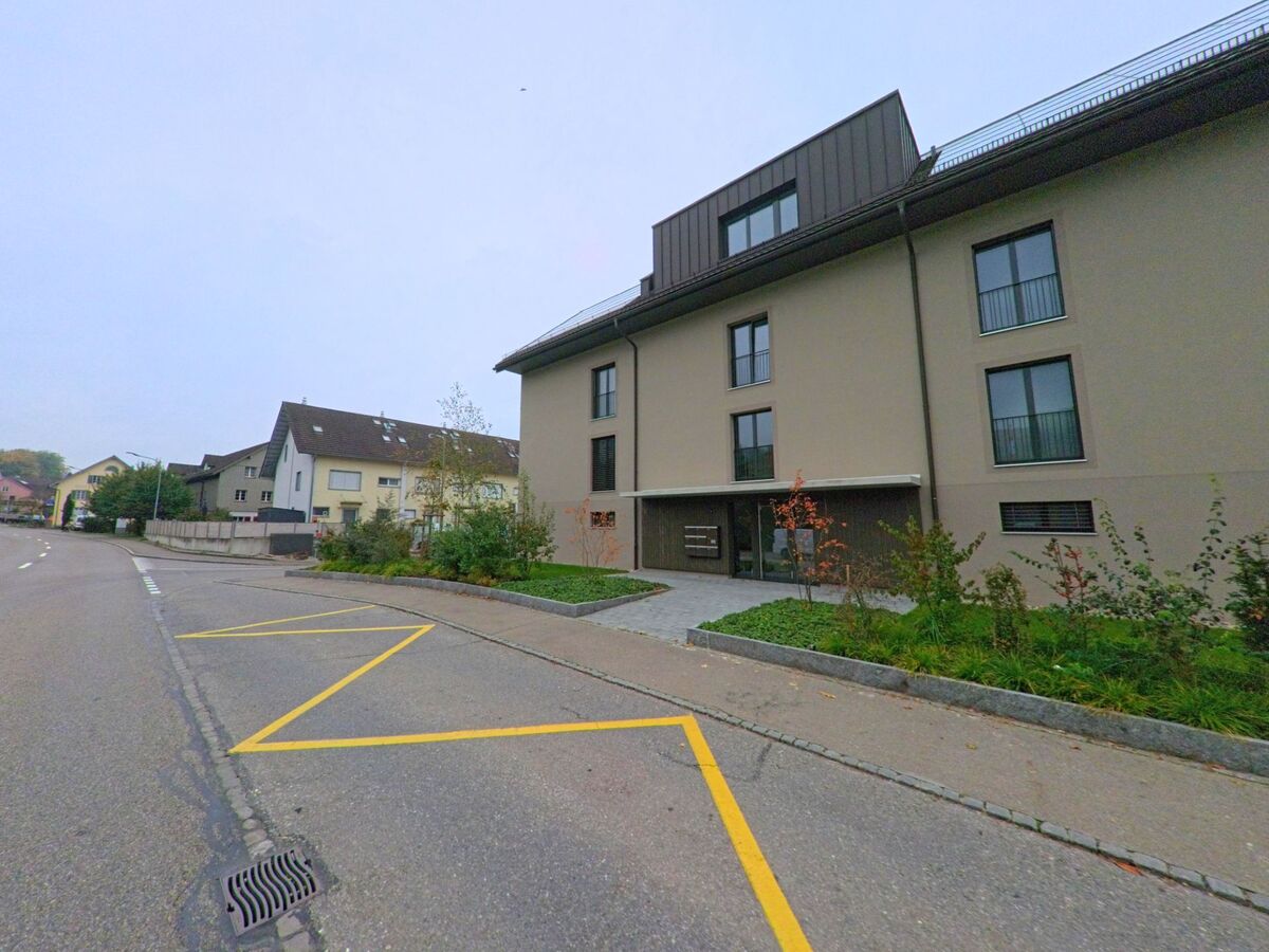 Ein modernes Wohnhaus in Künten (5444, Kanton Aargau) steht an einer Strasse mit markierten Parkplätzen und jungen Bäumen am Rand.