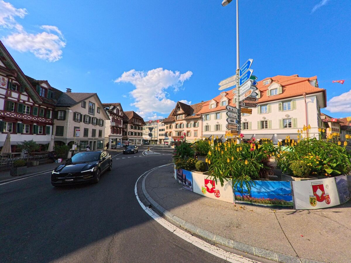 In Kuessnacht (6403, Kanton Schwyz) umrundet eine belebte Strasse einen bunt bepflanzten Kreisverkehr vor traditionellen Hausern mit Holz- und Putzfassaden.