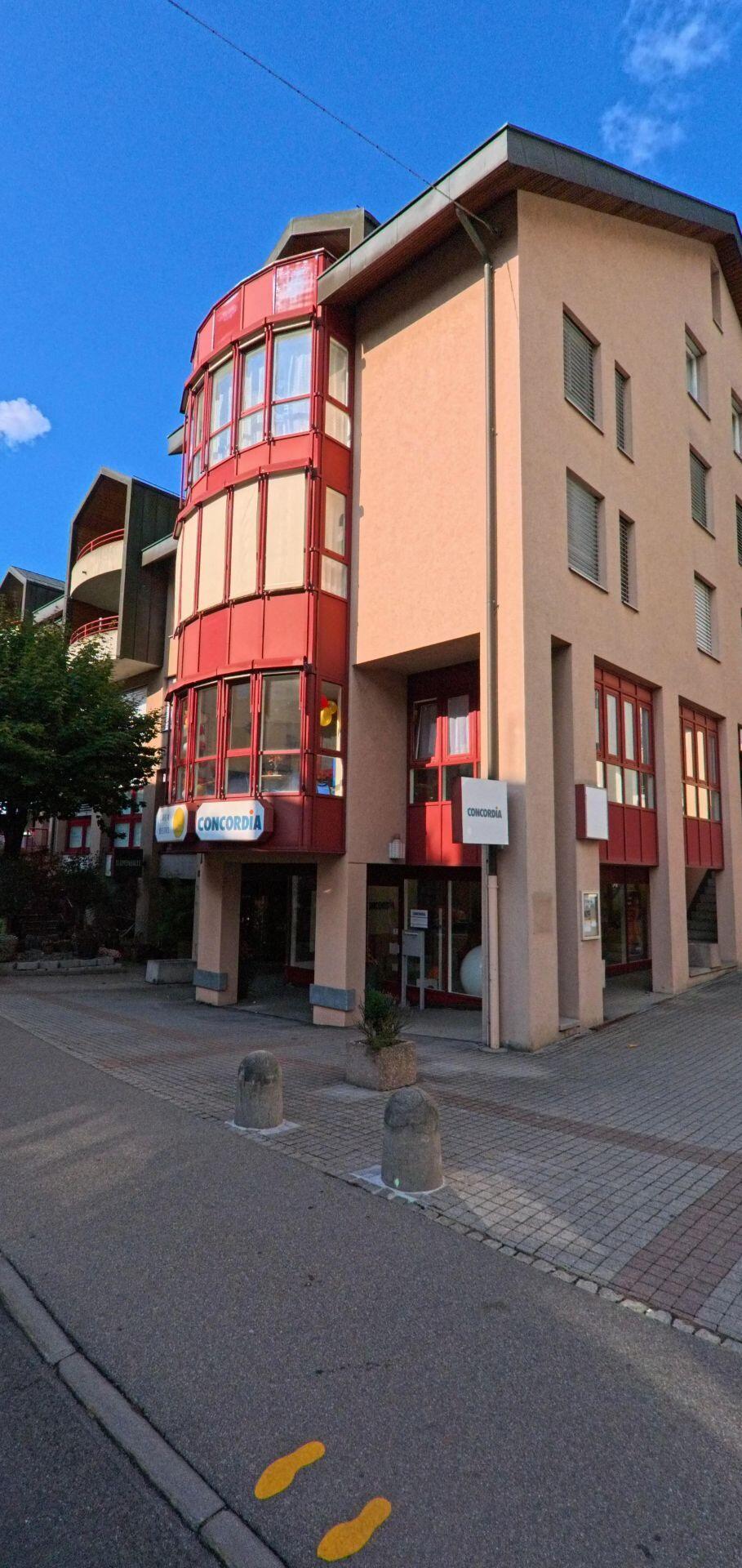 An einem modernen Gebaeude mit rot verglaster Rundfassade in Kuessnacht (6403, Kanton Schwyz) liegt ein kleiner Eingangsbereich mit Pollern und Pflastersteinen.