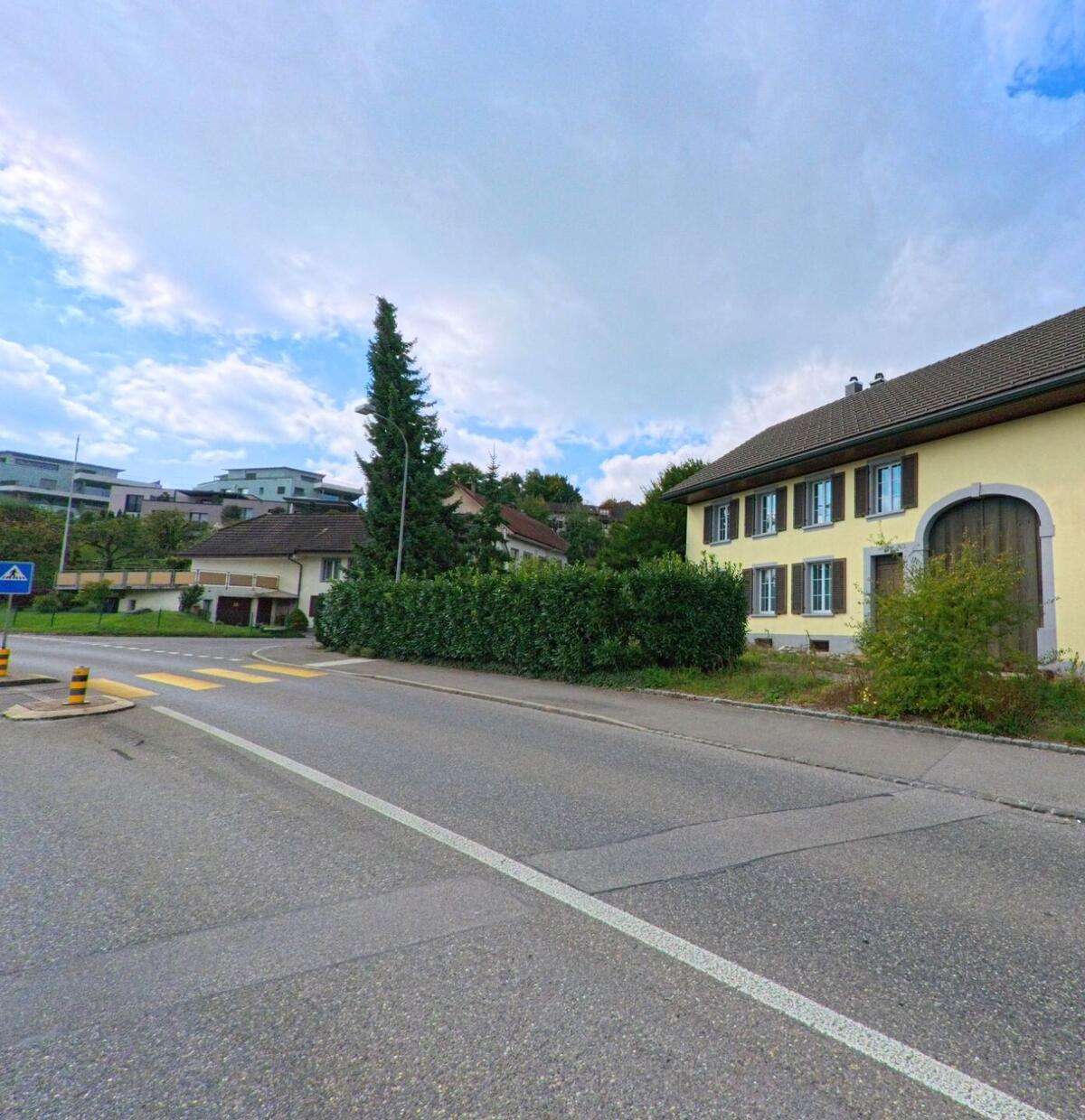 Ein gelbes Wohnhaus mit grossem Torbogen steht in Küttigen (5024, Kanton Aargau) an einer Strasse mit Fussgängerstreifen und Heckenbepflanzung.