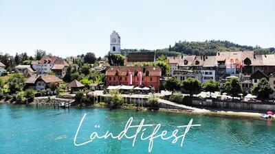 Am Ufer in Büren an der Aare (3294, Kanton Bern) zeigt das Bild das Ländtifest mit historischem Ortskern, Fahnen und Promenade entlang des türkisfarbenen Wassers.