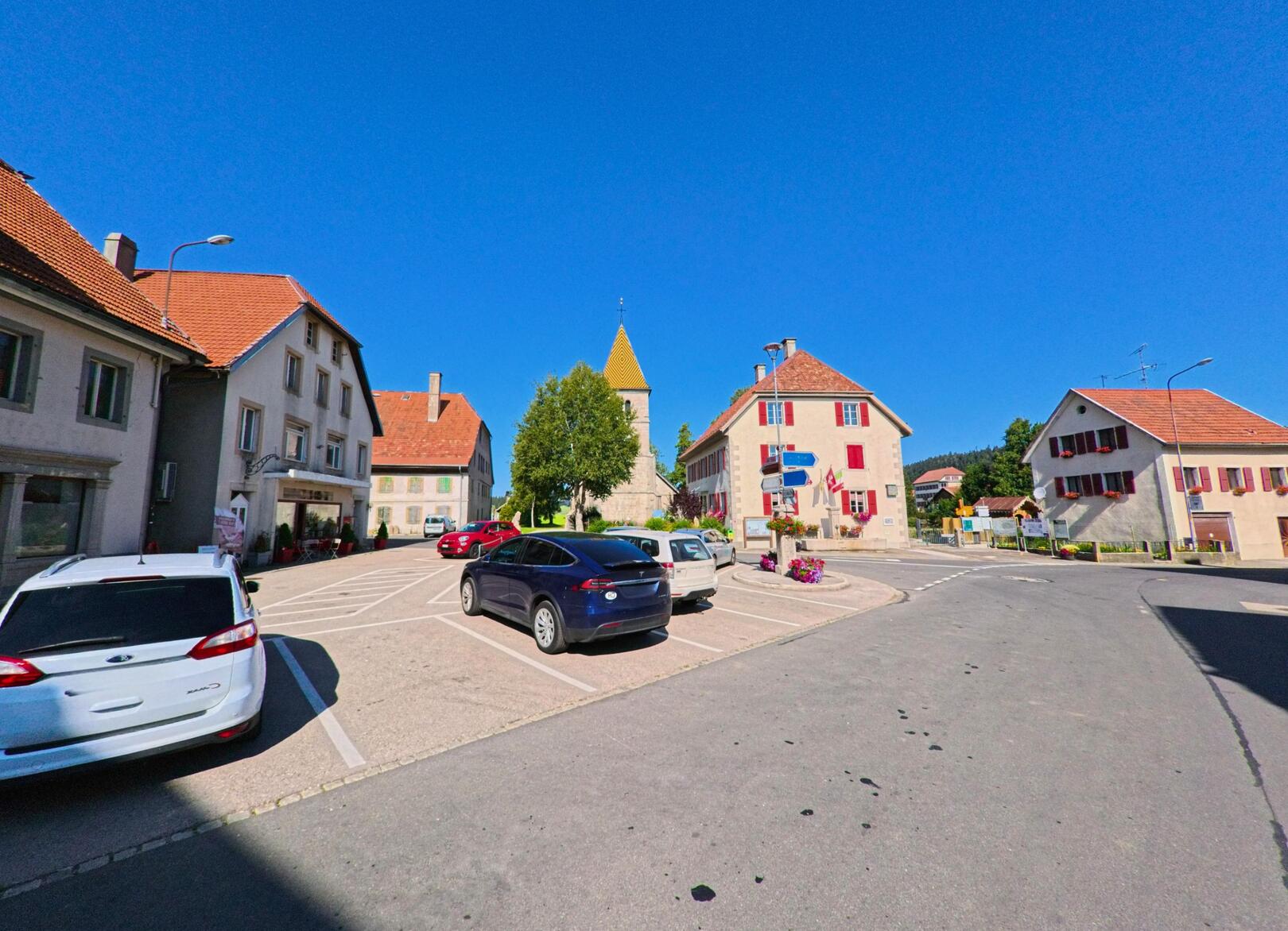Im Zentrum von La Brevine (2406, Kanton Neuenburg) stehen mehrere Gebaeude mit roten Ziegeldächern rund um einen Platz mit parkenden Autos, im Hintergrund ist der Kirchturm sichtbar.
