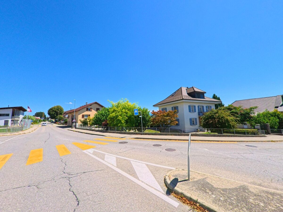 Entlang einer Hauptstrasse in La Brillaz (1745, Kanton Freiburg) stehen Wohnhaeuser mit gepflegten Gaerten, waehrend ein Fussgaengerstreifen die Fahrbahn markiert.