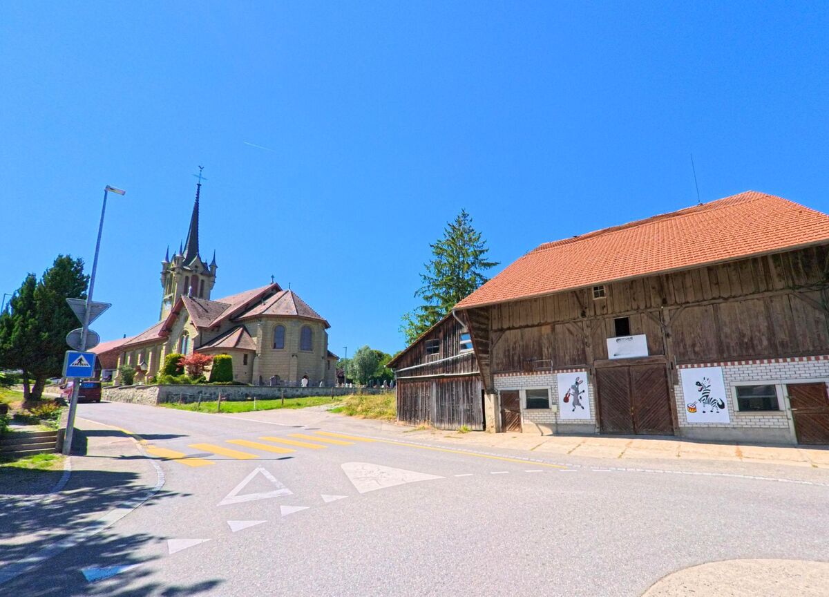 In La Brillaz (1756, Kanton Freiburg) steht eine Kirche mit hohem Turm neben einem grossen Holzgebaeude mit bemalten Tafeln an der Strassenkreuzung.