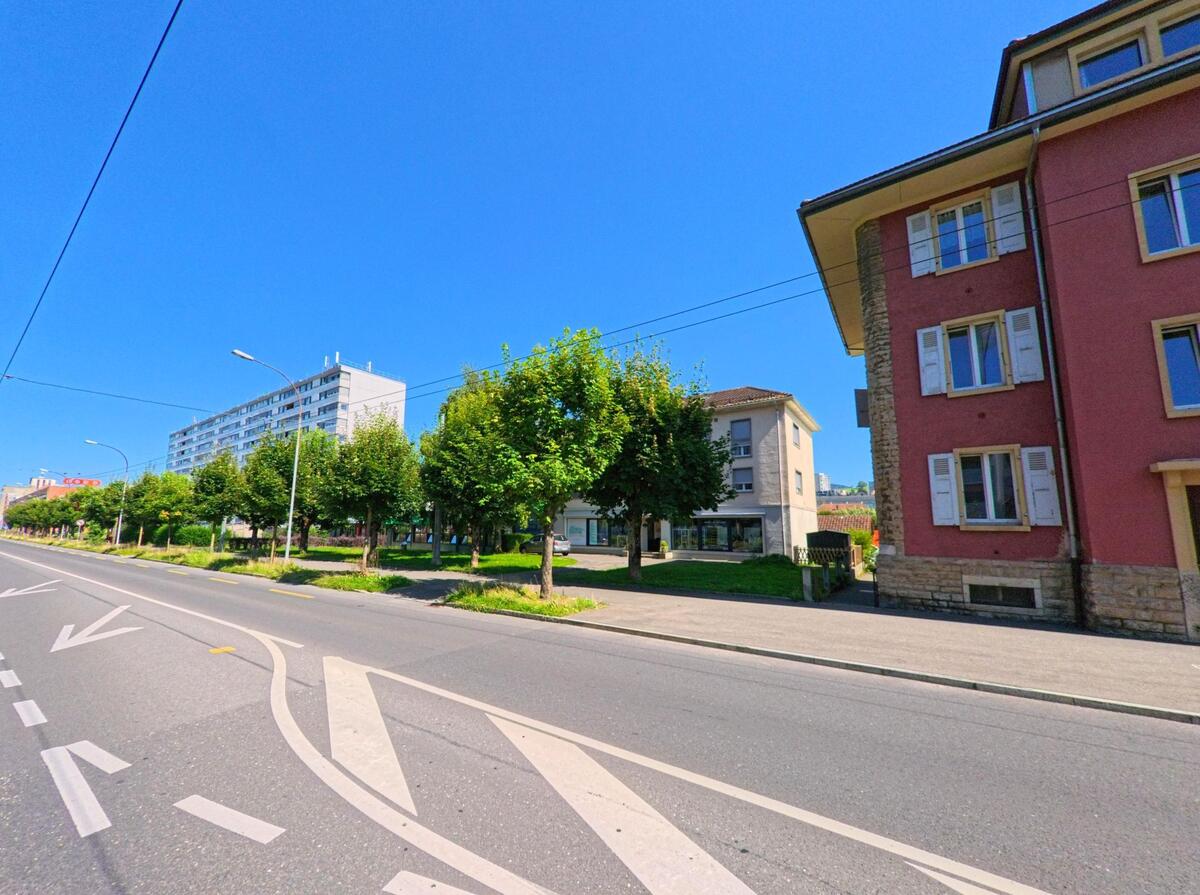 In La Chaux-de-Fonds (2300, Kanton Neuenburg) verlaeuft eine breite Strasse mit markierten Fahrspuren entlang von Wohnhaeusern und einer Reihe gepflegter Baeume.
