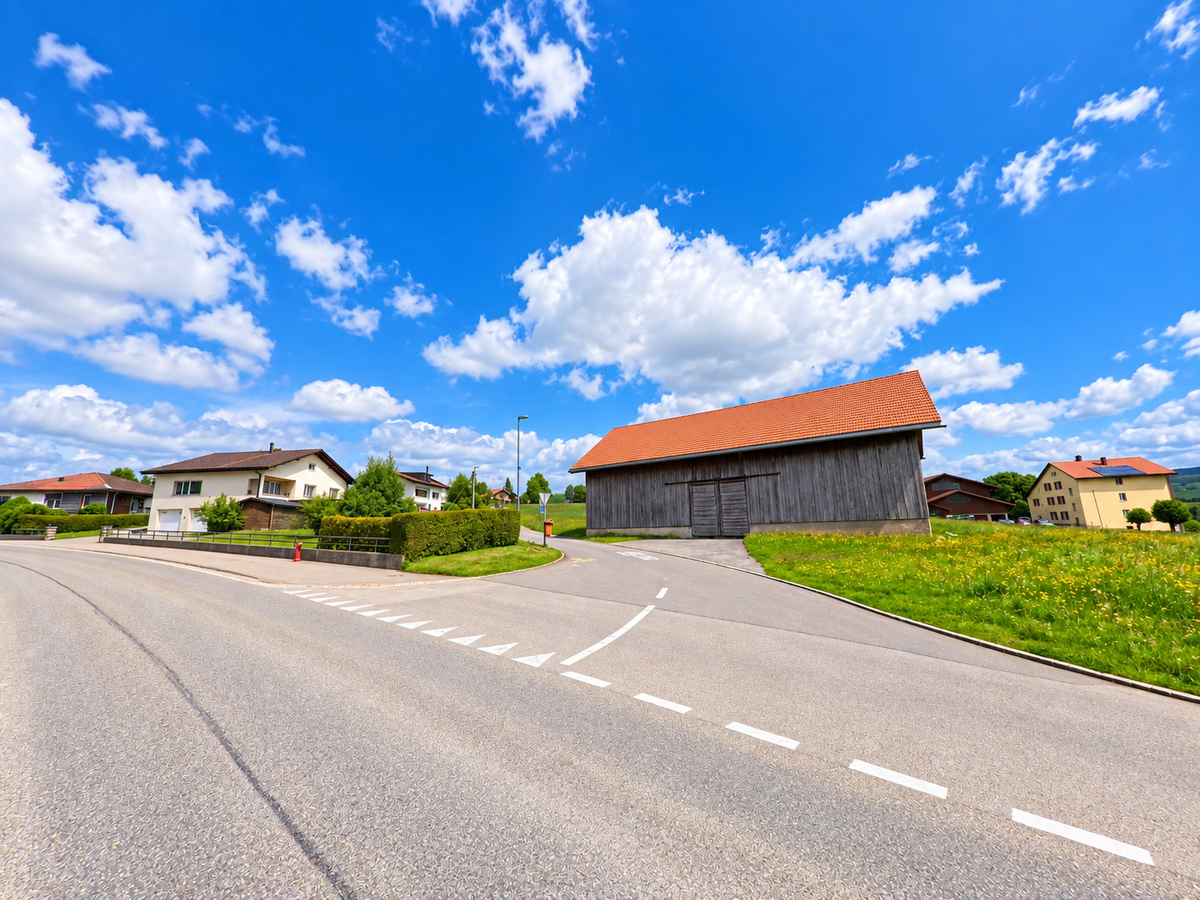 An einer Strassenverzweigung in La Chaux-des-Breuleux (2345, Kanton Jura) steht eine grosse Holzscheune zwischen Wohnhäusern und blühenden Wiesen.
