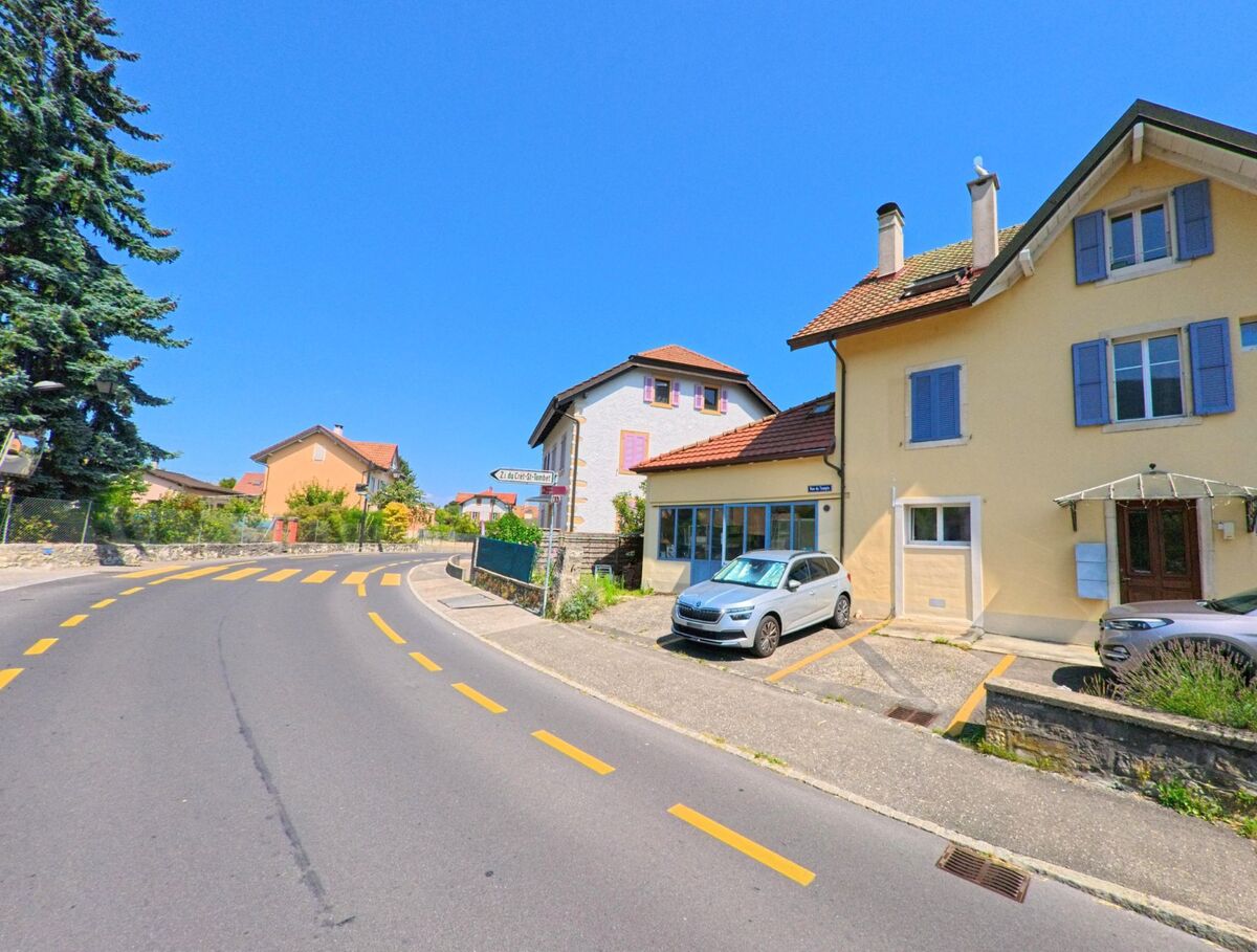 In La Grande Beroche (2022, Kanton Neuenburg) fuehrt eine leicht geschwungene Hauptstrasse an Wohnhaeusern mit hellen Fassaden vorbei.