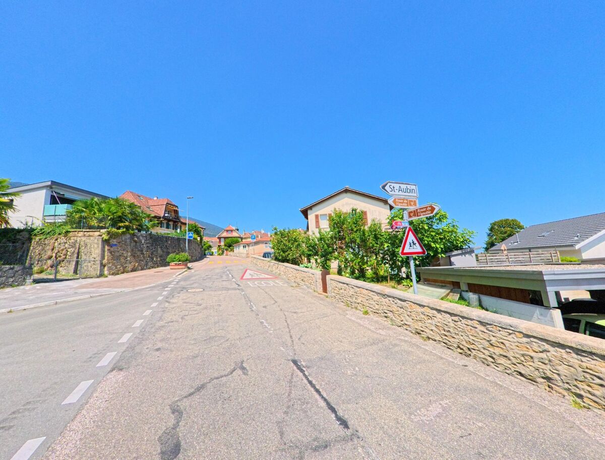 Eine ansteigende Strasse in La Grande Beroche (2023, Kanton Neuenburg) fuehrt zwischen Wohnhaeusern bergauf, waehrend Wegweiser unter anderem nach St-Aubin zeigen.