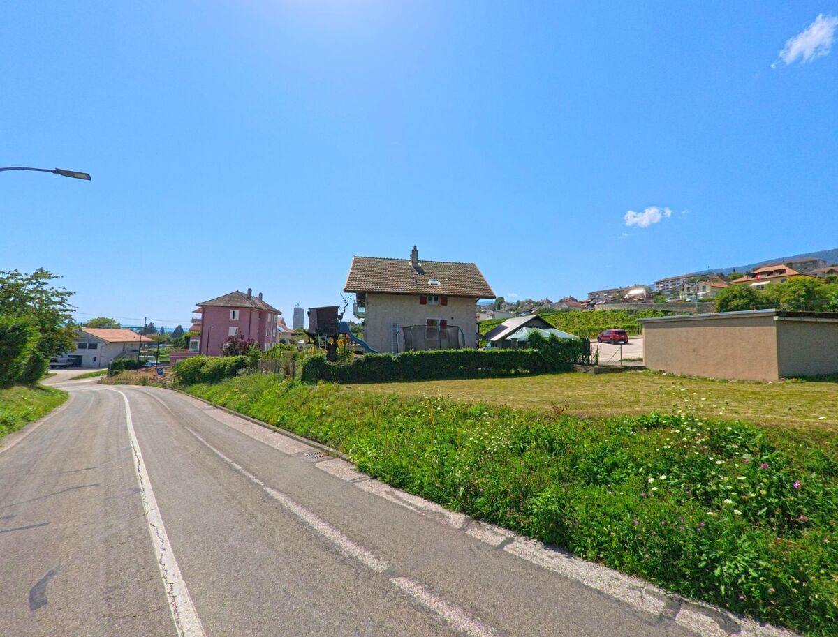 Eine Landstrasse in La Grande Beroche (2023, Kanton Neuenburg) fuehrt an Einfamilienhaeusern vorbei, waehrend im Hintergrund weitere Haeuser am Hang sichtbar sind.