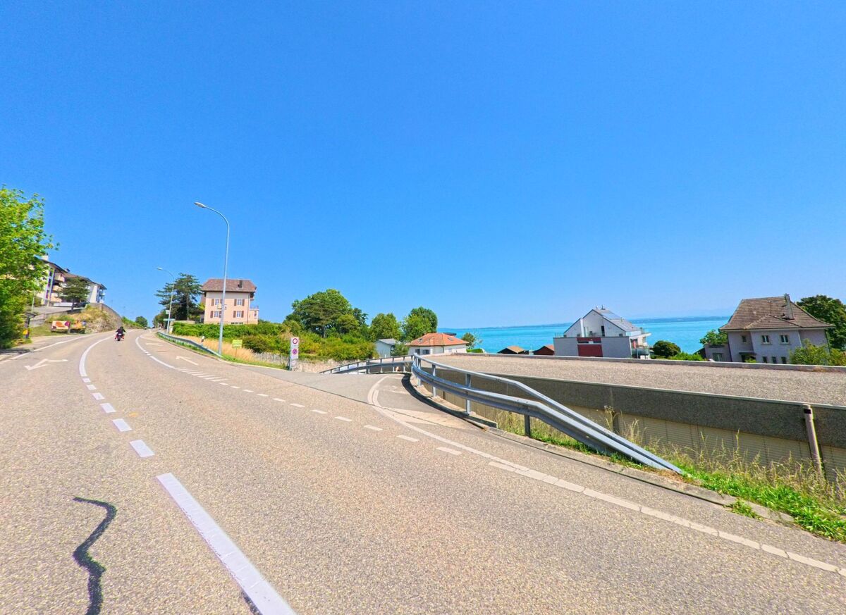 Eine Strasse in La Grande Beroche (2025, Kanton Neuenburg) fuehrt bergauf und gibt zugleich den Blick auf den Neuenburgersee und umliegende Haeuser frei.