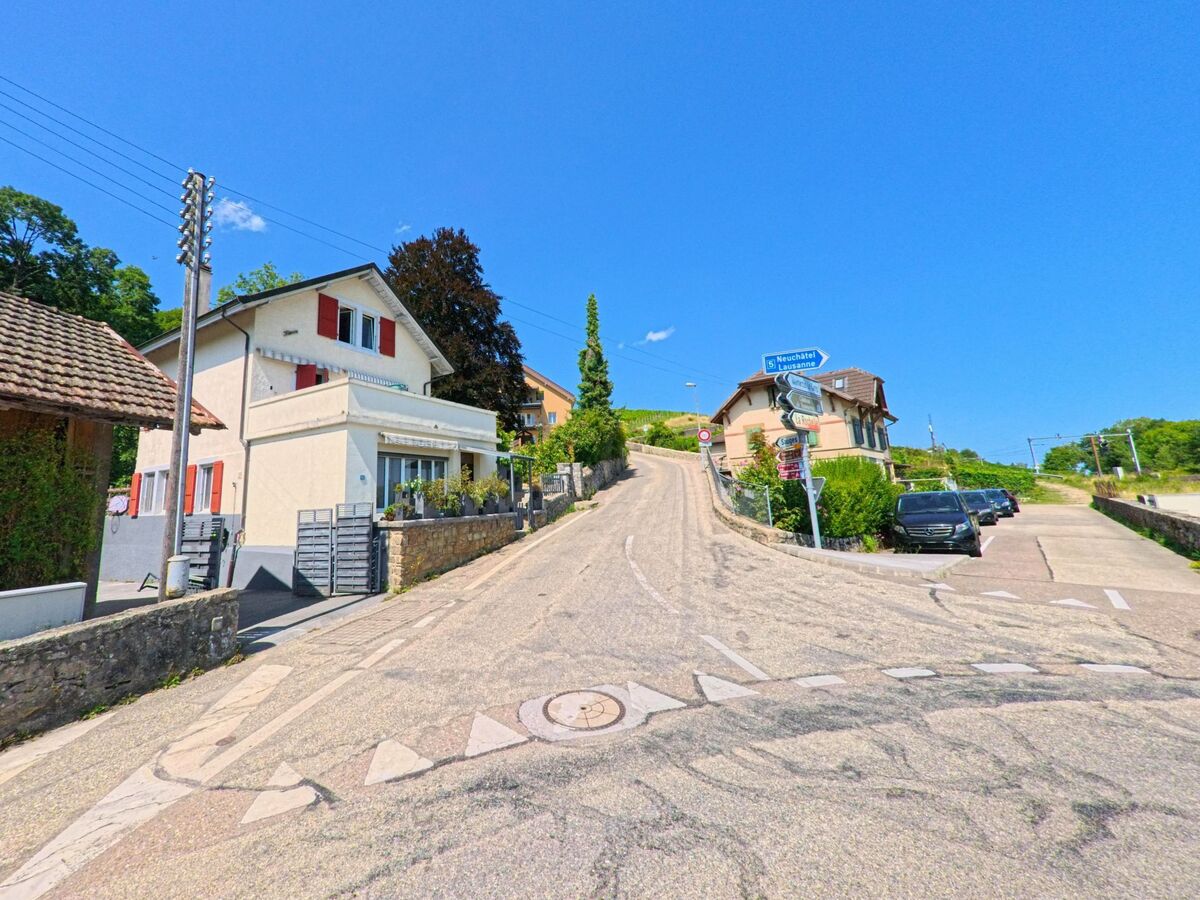 Zwischen Wohnhaeusern in La Grande Beroche (2028, Kanton Neuenburg) verlaeuft eine ansteigende Strasse mit Wegweisern in Richtung Neuchatel und Vaumarcus.