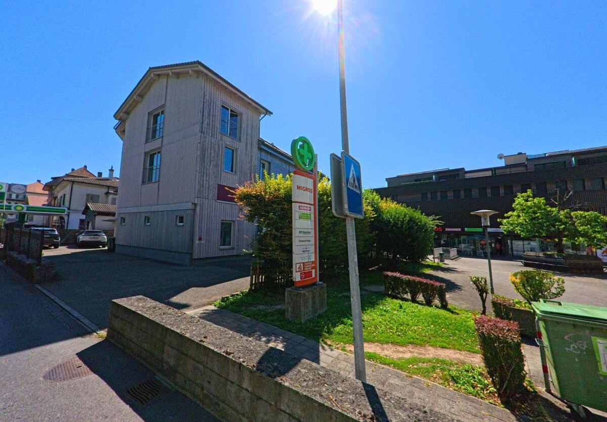 In La Neuveville (2520, Kanton Bern) steht ein moderner Gebaeudekomplex mit Einkaufsmoeglichkeiten neben einem Strassenschild und einer Bushaltestelle.