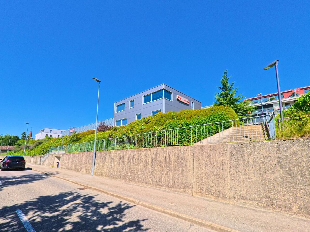 Ein modernes Gewerbegebaeude auf einer Anhhoe in La Neuveville (2520, Kanton Bern) wird von einer Treppe und gruener Bepflanzung eingerahmt.