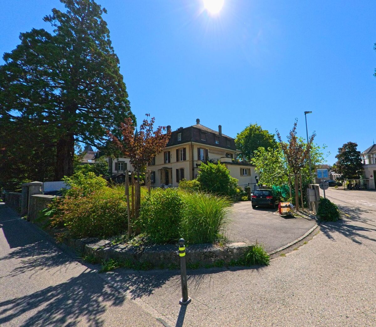 Vor einem grossen Nadelbaum steht ein elegantes Wohnhaus mit Garten in La Neuveville (2520, Kanton Bern) bei strahlendem Sonnenschein.