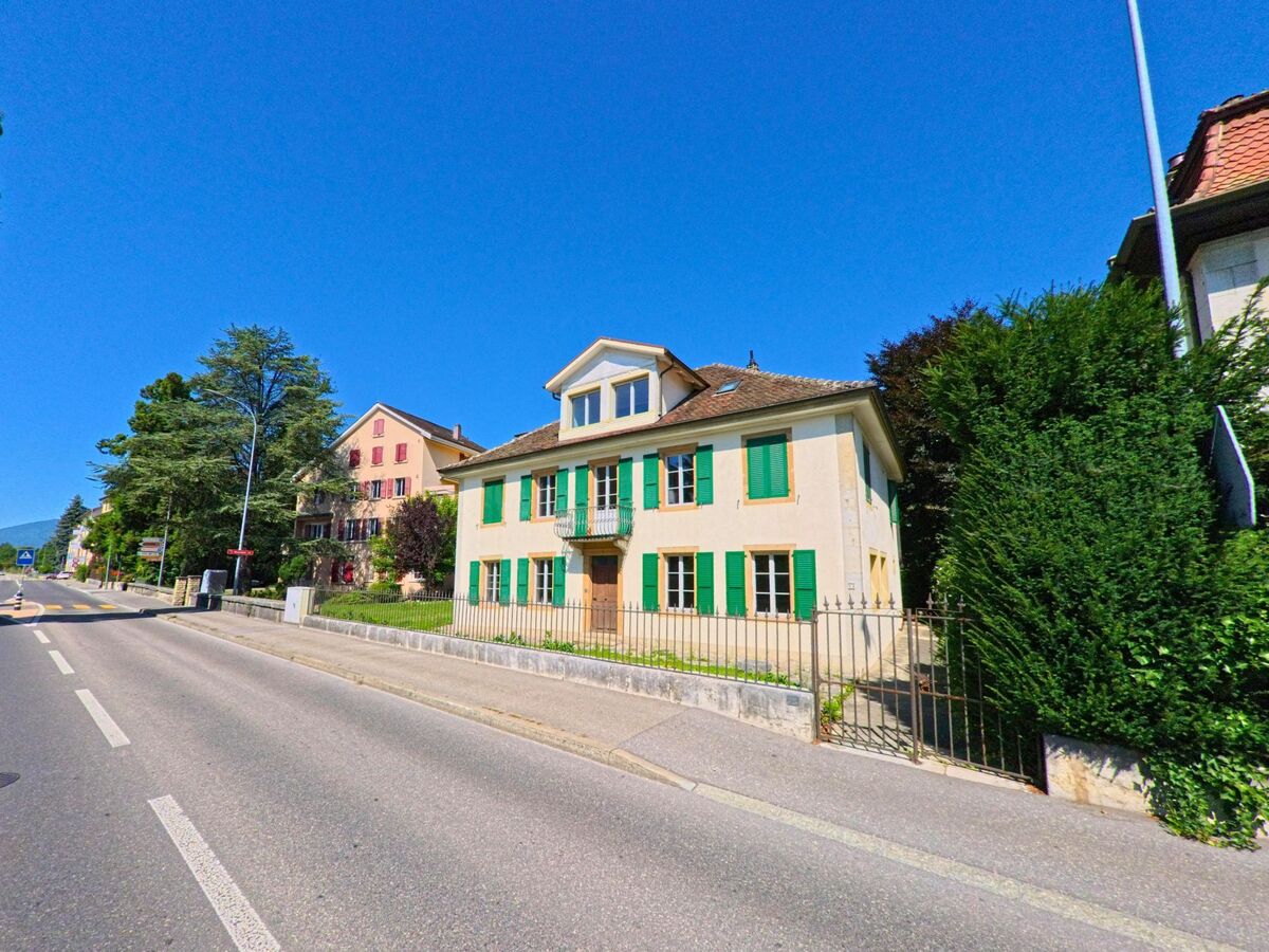 Ein helles Wohnhaus mit gruenen Fensterlaeden steht an einer Hauptstrasse in La Neuveville (2520, Kanton Bern) unter blauem Himmel.