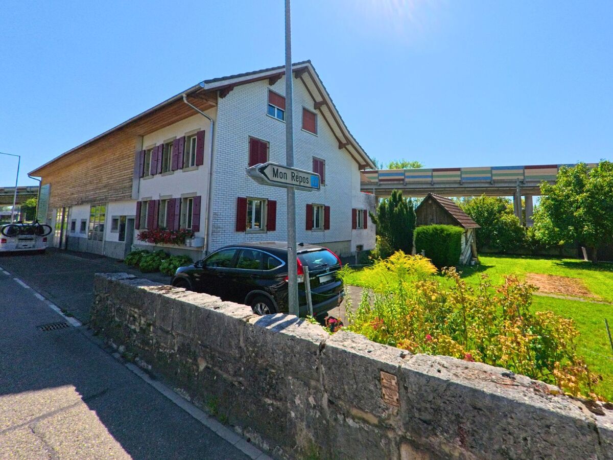 Ein Wohnhaus mit weisser Fassade und roten Fensterlaeden steht in La Neuveville (2520, Kanton Bern) nahe einer farbigen Brueckenanlage.