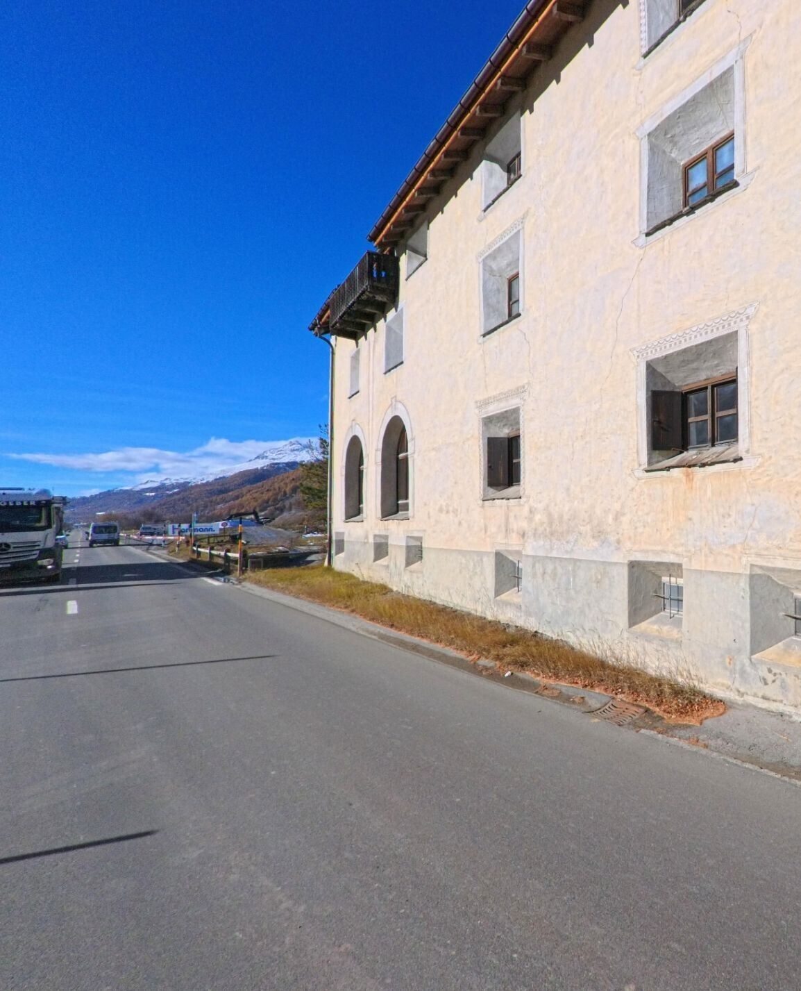 Entlang einer Hauptstrasse verläuft die Fassade eines historischen Gebäudes am Rand von La Punt Chamues-ch (7522, Kanton Graubünden), während sich dahinter die alpine Landschaft öffnet.
