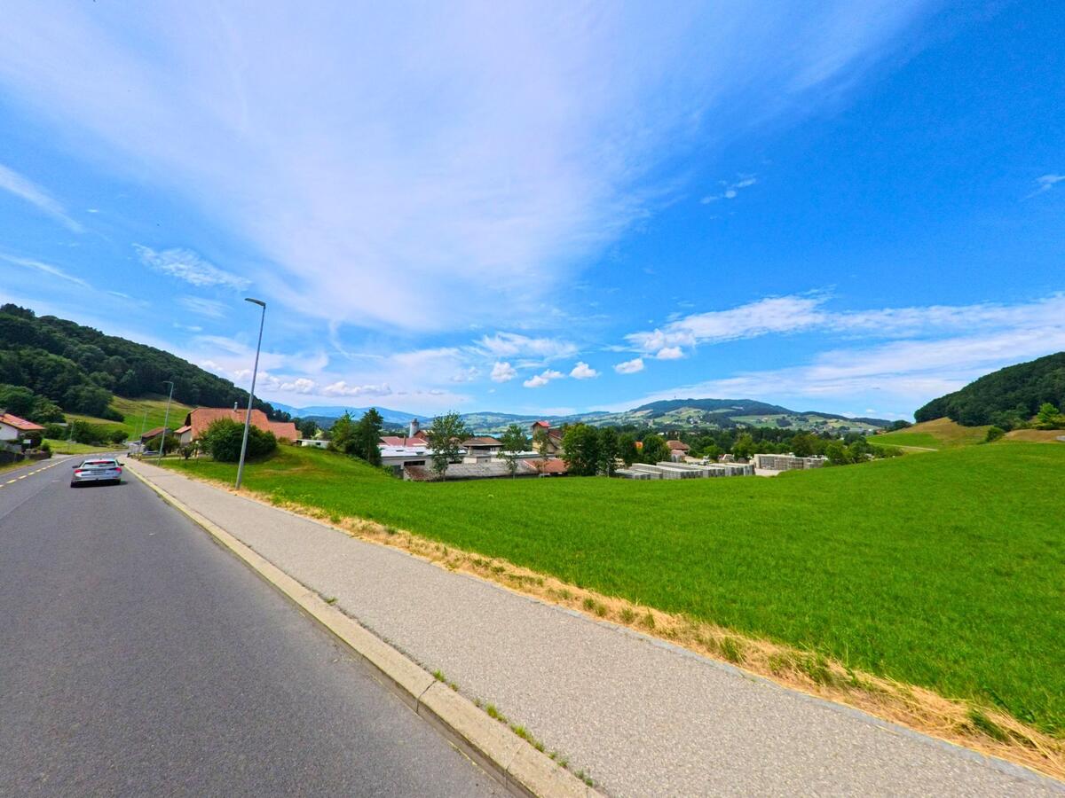In La Roche (1634, Kanton Freiburg) führt eine Strasse mit Trottoir an weitläufigen Wiesen vorbei auf das Dorfzentrum mit Blick auf die umliegenden Hügel zu.