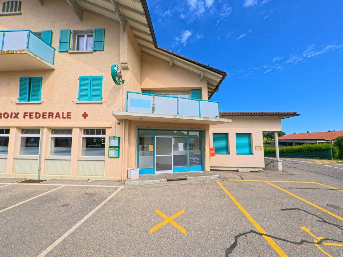 Vor einem Gebäude mit auffälligen blauen Fensterläden in La Verrerie (1624, Kanton Freiburg) befinden sich markierte Parkplätze und ein Eingang mit angeschlossenem Postfachbereich.