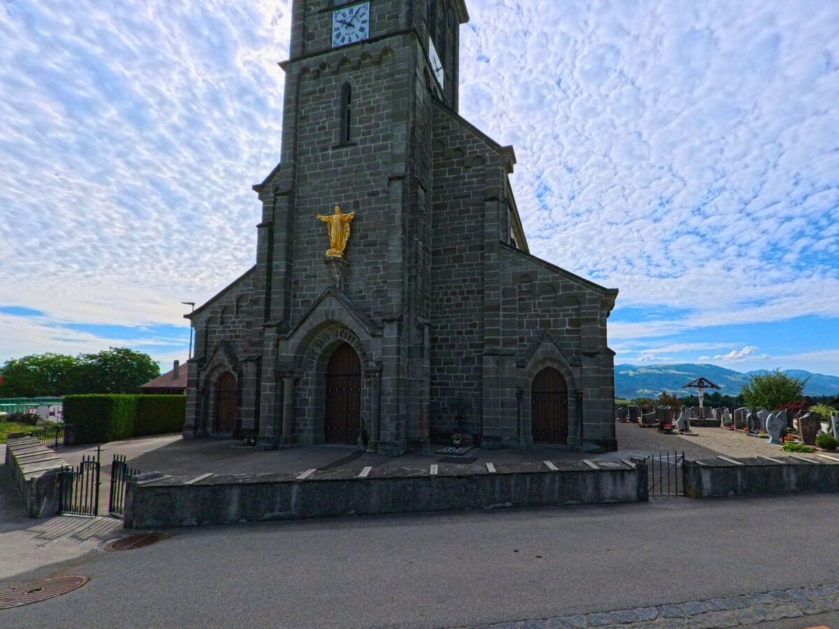 Die Kirche von La Verrerie (1624, Kanton Freiburg) mit markanter Christusstatue am Turm liegt direkt neben einem gepflegten Friedhof mit Blick auf die umliegenden Hügel.