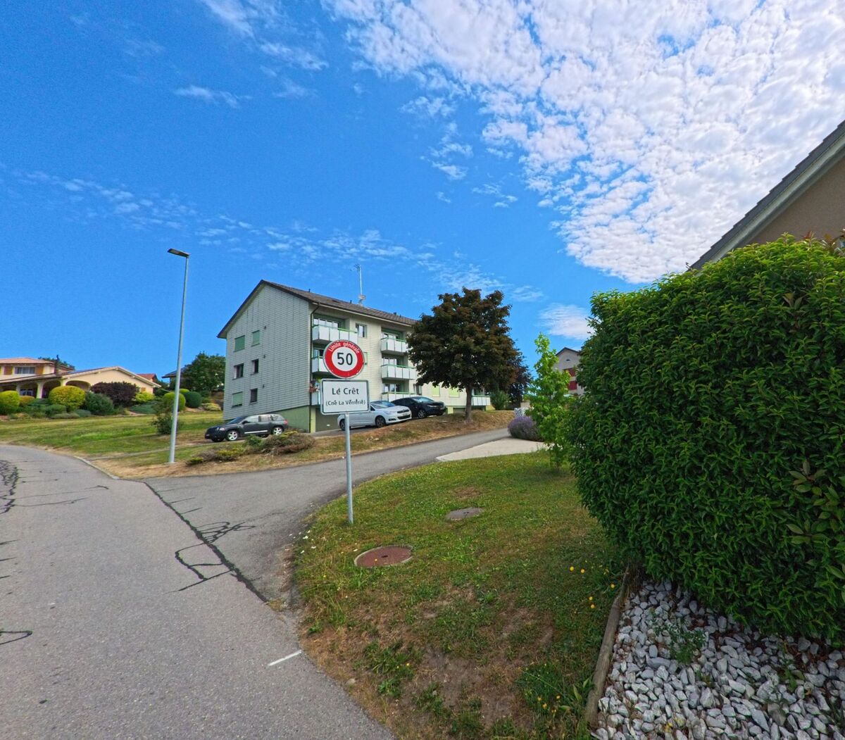 Am Ortseingang des Ortsteils Le Crêt in La Verrerie (1624, Kanton Freiburg) steht ein Geschwindigkeitsschild vor einem Wohnquartier mit Mehrfamilienhäusern und gepflegten Gärten.