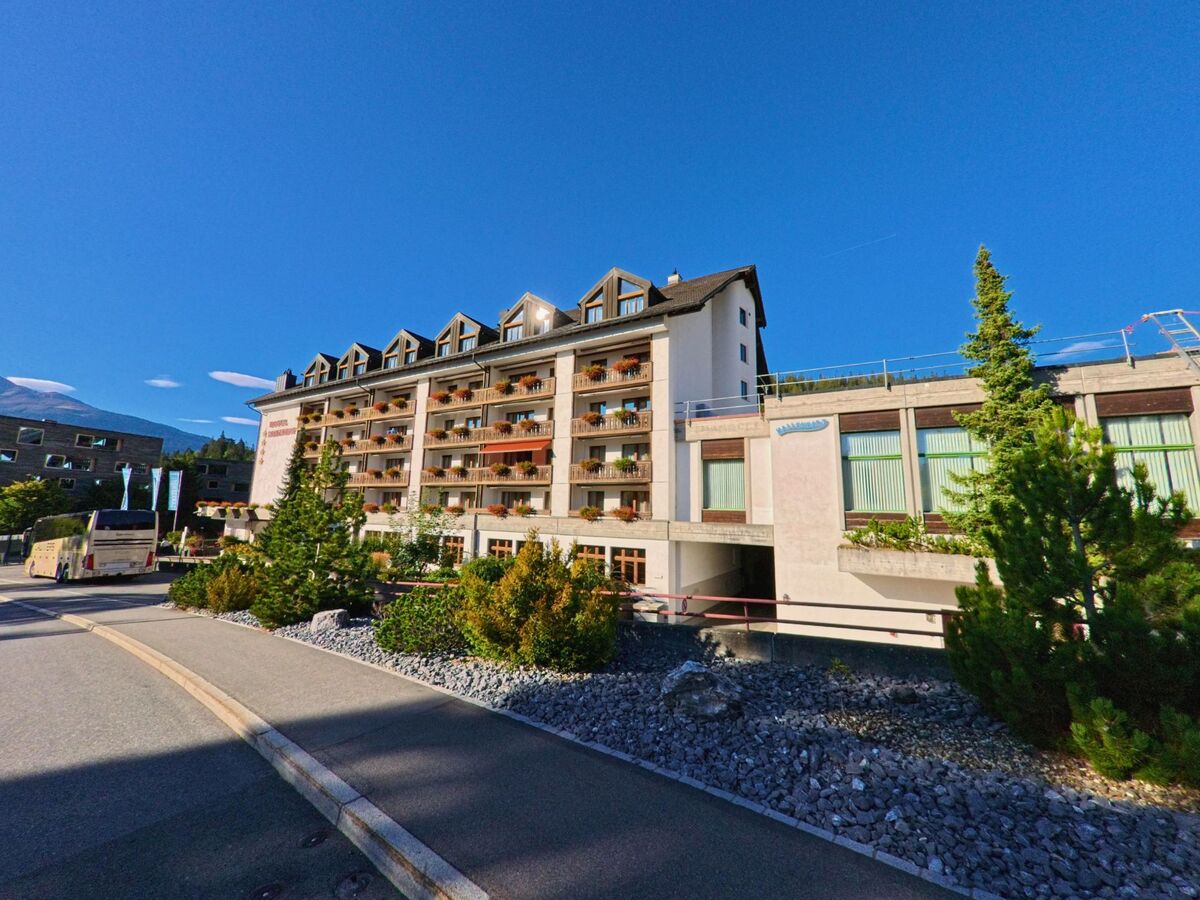 Das Hotel Laaxerhof in Laax (7032, Kanton Graubuenden) zeigt seine sonnenbeschienene Fassade mit Blumenkästen und angrenzender Strasse vor blauem Himmel.