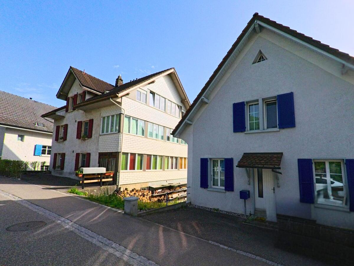 Zwei Einfamilienhäuser mit farbigen Fensterläden prägen das ruhige Strassenbild in Läufelfingen (4448, Kanton Baselland) bei Sonnenschein.