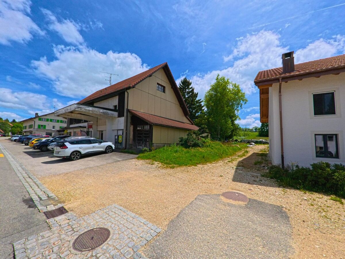 In Lajoux (2718, Kanton Jura) steht ein älteres Wirtschaftsgebäude mit überdachtem Vorbau neben einer Reihe geparkter Fahrzeuge und einem Schotterweg ins Grüne.
