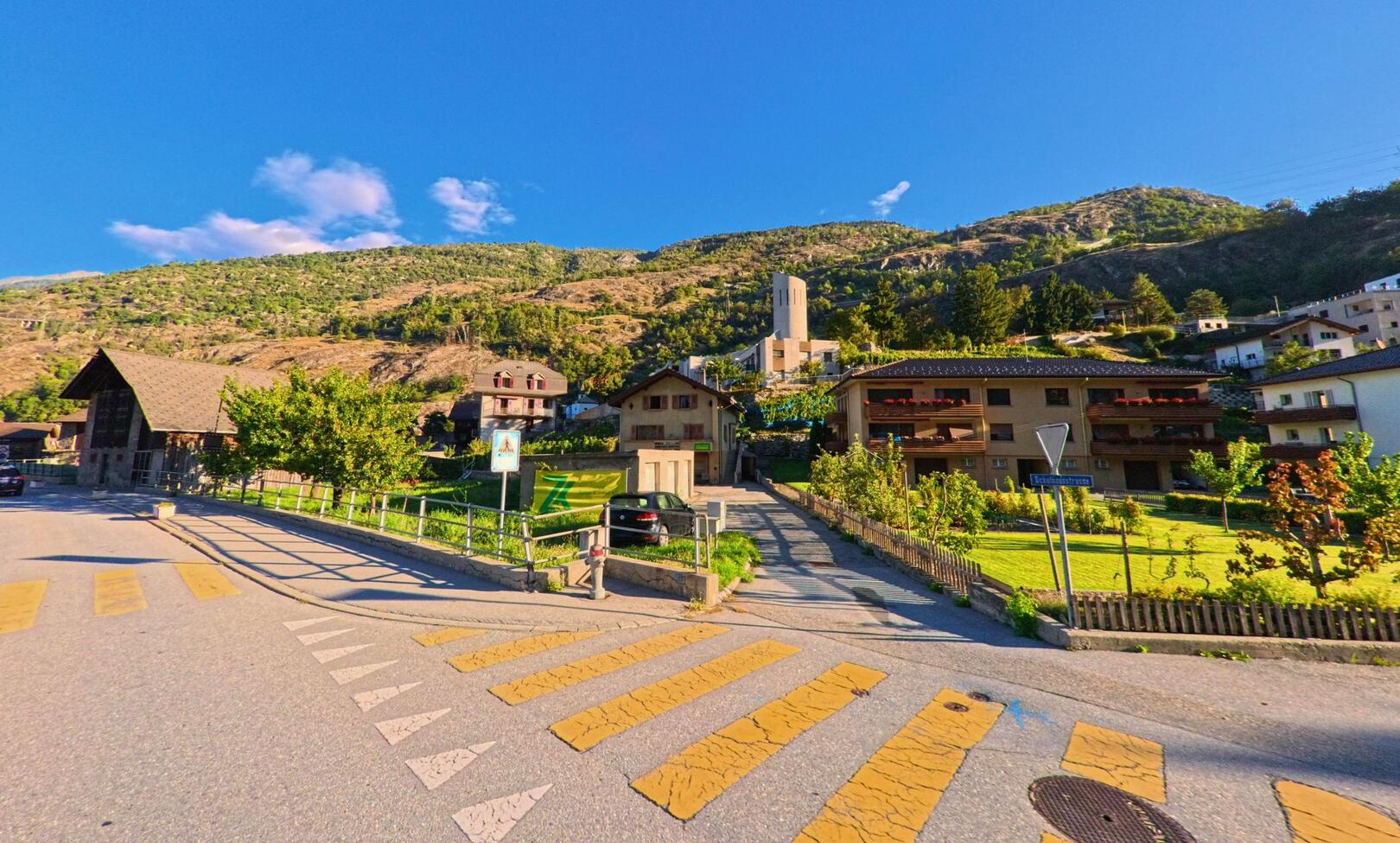 Ein Fussgaengerstreifen fuehrt in Lalden (3931, Kanton Wallis) auf eine Strasse mit Wohnhaeusern und Kirche, im Hintergrund erheben sich bewaldete Huegel.