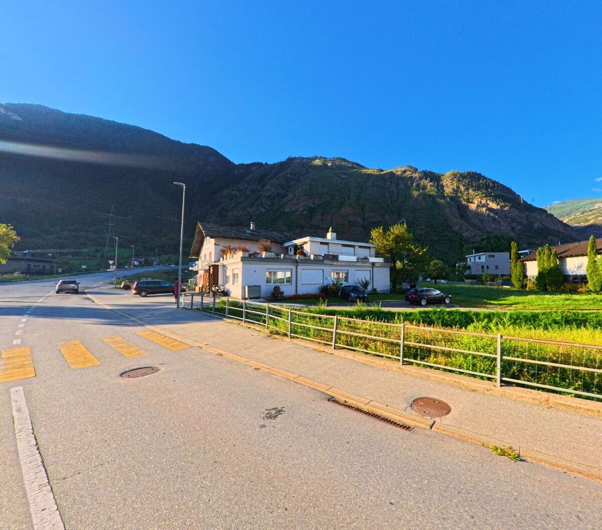 Ein Fussgaengerstreifen fuehrt in Lalden (3931, Kanton Wallis) an einer Strasse vorbei, neben der einzelne Wohnhaeuser und Felder am Fusse der Berge liegen.