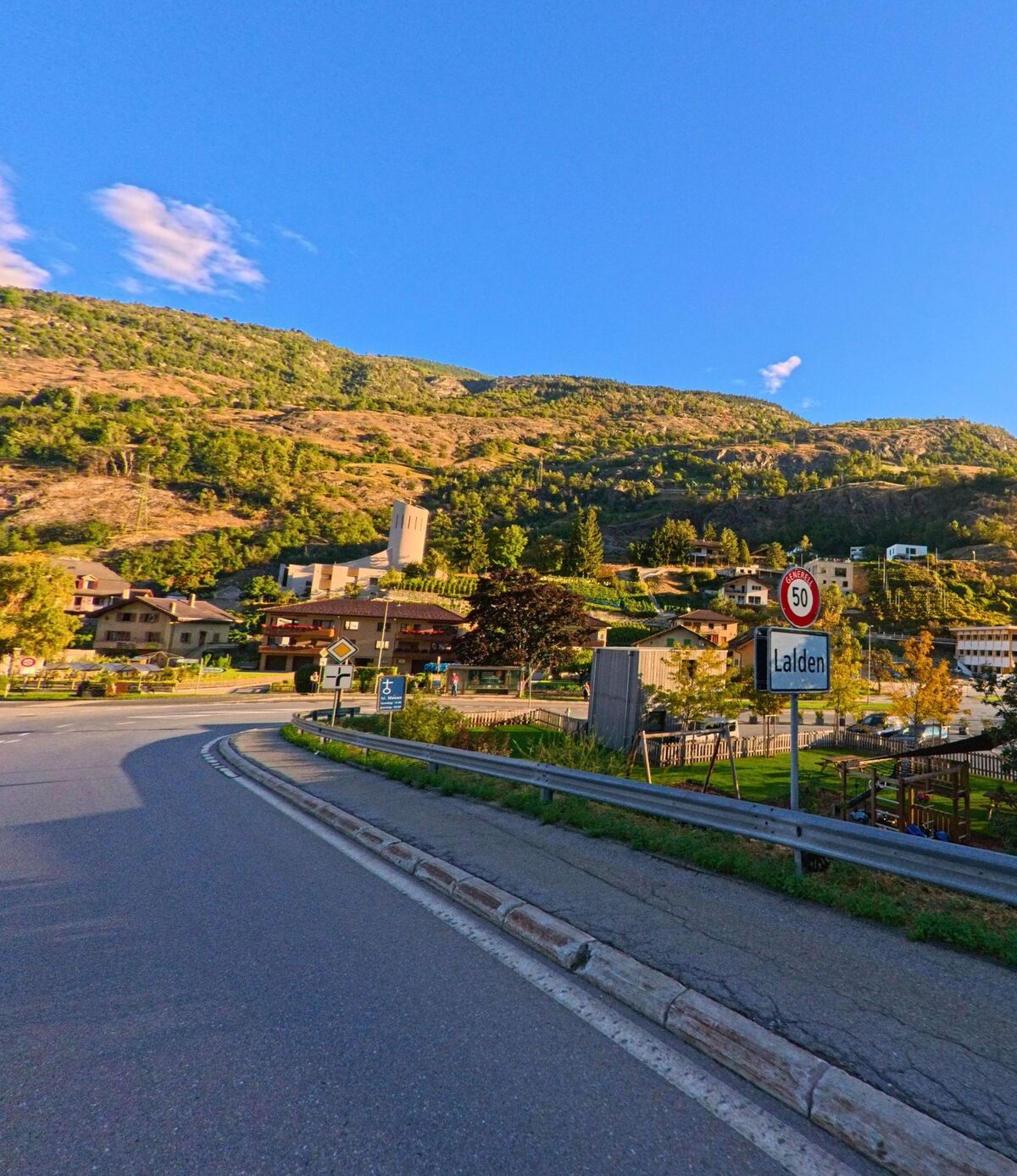 An der Ortseinfahrt von Lalden (3931, Kanton Wallis) steht ein Verkehrsschild mit Geschwindigkeitsbegrenzung, im Hintergrund sind Kirche und Wohnhaeuser am Hang sichtbar.