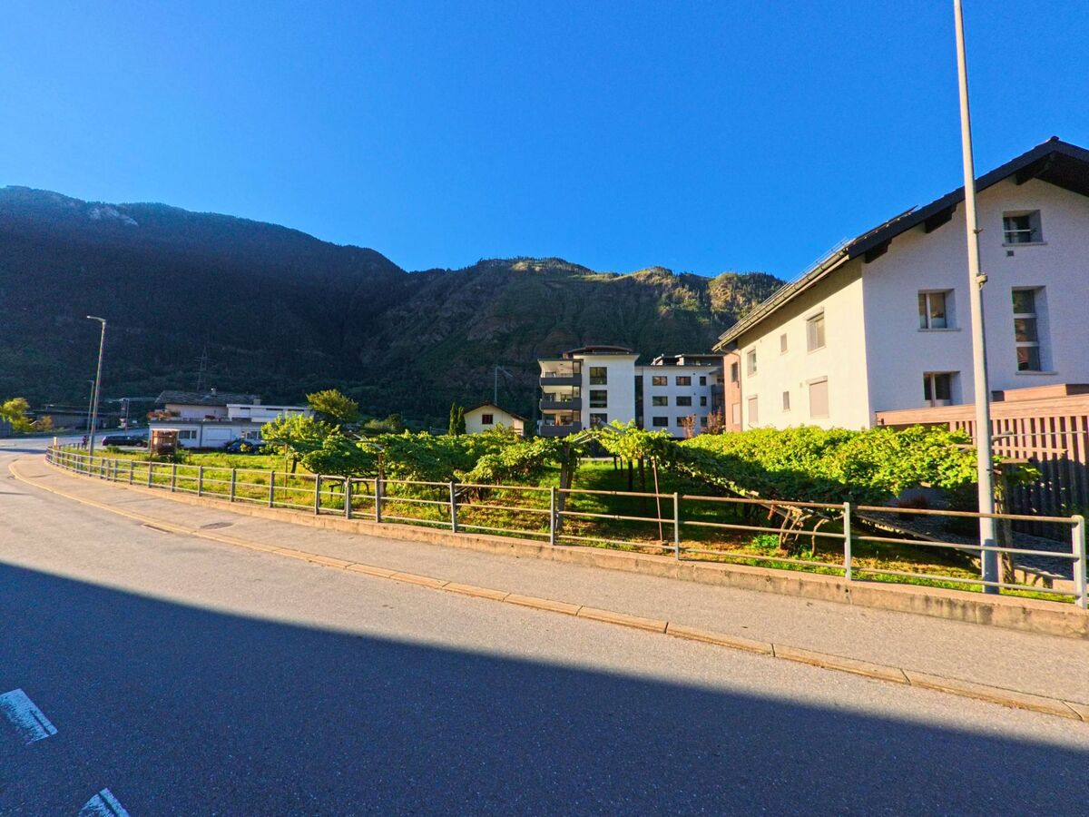 Zwischen Wohnhaeusern und einer kleinen Rebparzelle in Lalden (3931, Kanton Wallis) fuehrt die Strasse in einer Kurve am Fuss der bewaldeten Berge entlang.