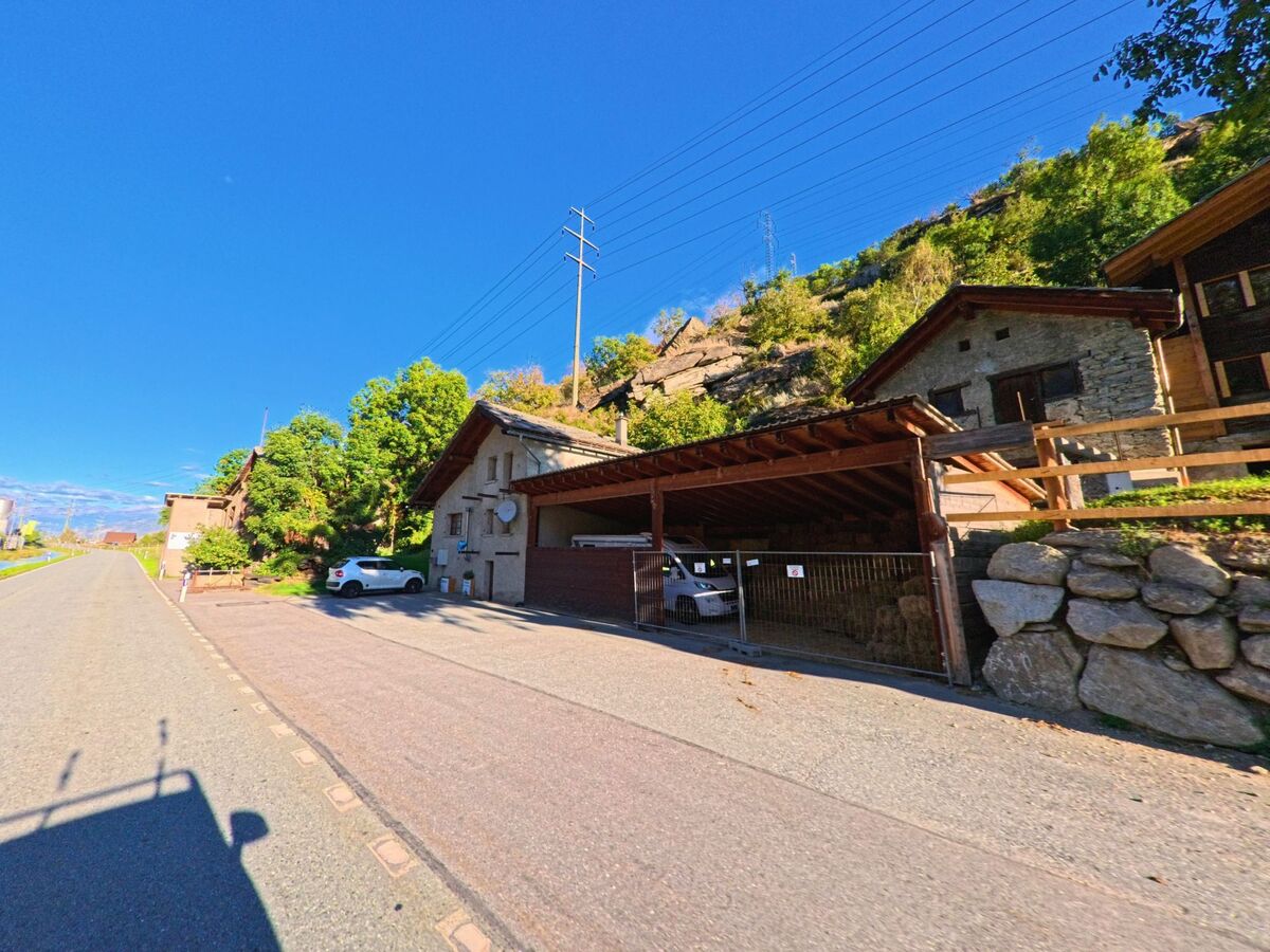 Am Hang in Lalden (3931, Kanton Wallis) stehen traditionelle Steinhaeuser und ein Carport, daneben verlaeuft eine ruhige Strasse unter blauem Himmel.