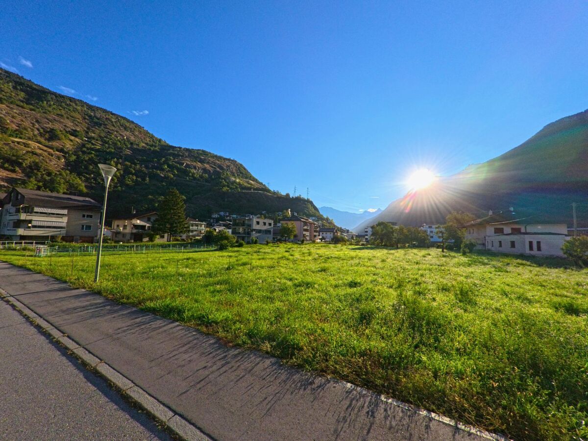 Eine gruene Wiese erstreckt sich in Lalden (3931, Kanton Wallis) am Dorfrand, waehrend die Sonne zwischen den Bergen untergeht und die Haeuser im Hintergrund beleuchtet.