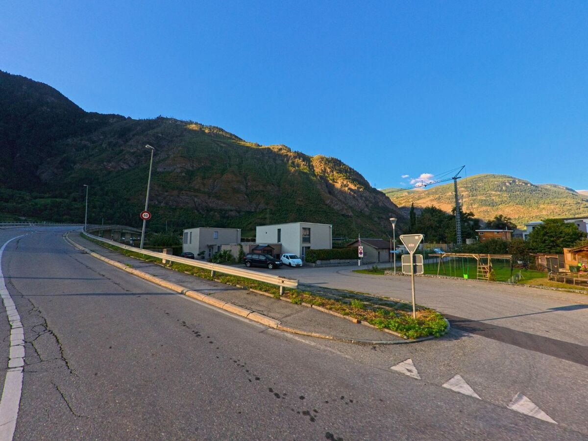 Eine ansteigende Strasse fuehrt in Lalden (3931, Kanton Wallis) an modernen Wohnhaeusern vorbei, waehrend im Hintergrund Baukraene und bewaldete Berge zu sehen sind.