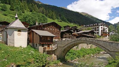 Im Dorf Binn (3996, Kanton Wallis) im Landschaftspark Binntal überspannt eine alte Steinbrücke den Bach, umgeben von traditionellen Holzhäusern und grünen Berghängen.