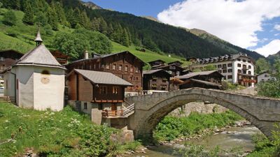 Im Dorf Binn (3996, Kanton Wallis) im Landschaftspark Binntal überspannt eine alte Steinbrücke den Bach, umgeben von traditionellen Holzhäusern und grünen Berghängen.