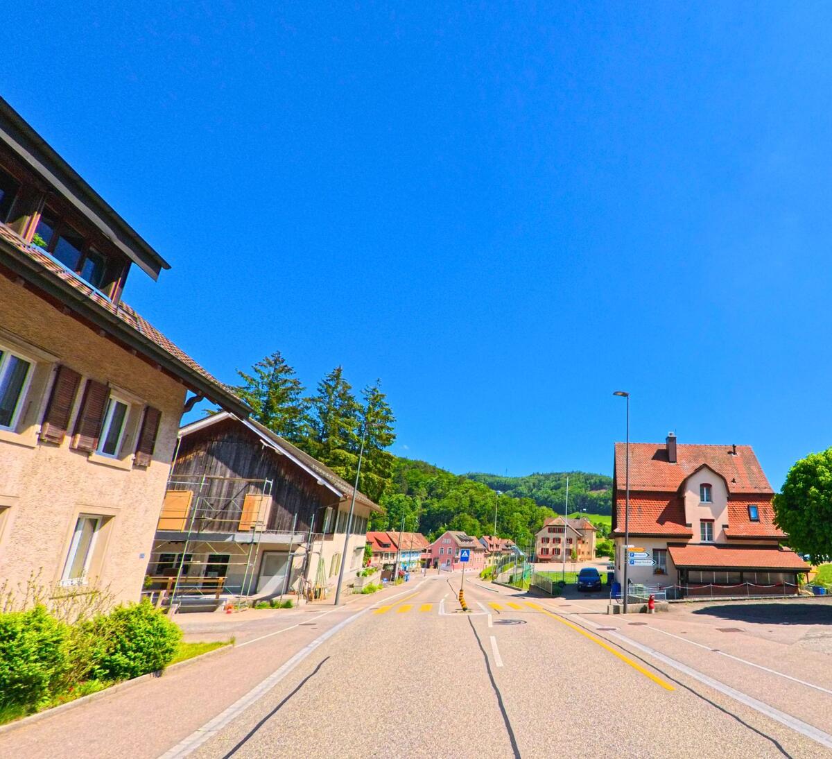 Im unteren Bereich von Langenbruck (4438, Kanton Baselland) öffnet sich die Strasse zu einem kleinen Platz mit Parkmöglichkeiten und umstehenden Wohnhäusern vor bewaldeten Hügeln.