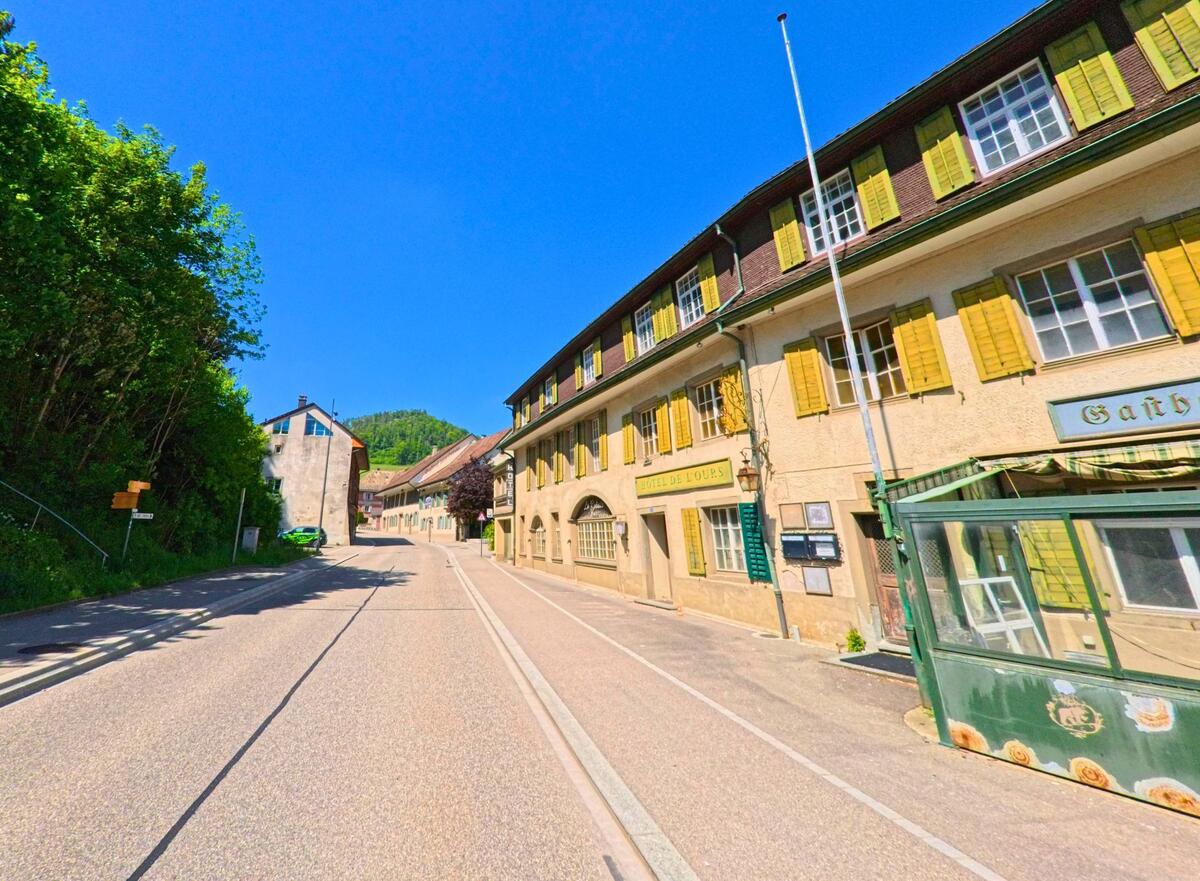 In Langenbruck (4438, Kanton Baselland) liegt das historische Hotel de l’Ours mit gelben Fensterläden direkt an der Hauptstrasse, gegenüber eines bewaldeten Hangs.