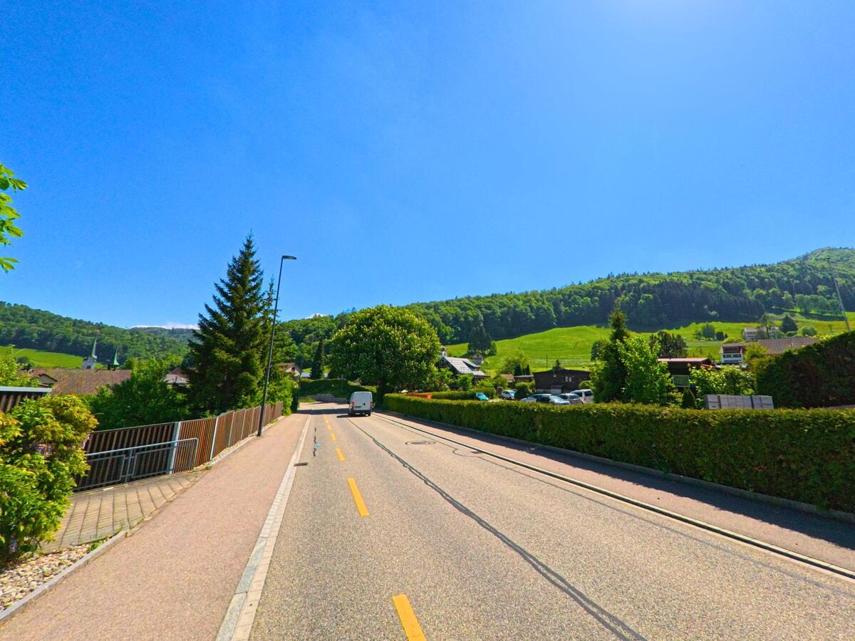 Eine leicht ansteigende Strasse in Langenbruck (4438, Kanton Baselland) führt durch ein ruhiges Wohnquartier mit Blick auf bewaldete Hügel und grüne Wiesen.