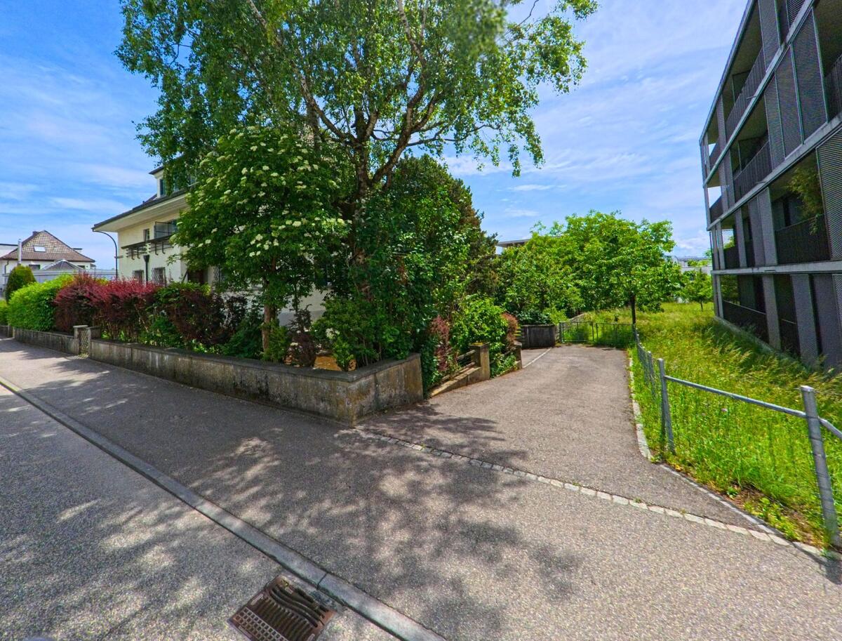 In Langendorf (4513, Kanton Solothurn) zweigt ein schattiger Fussweg zwischen modernen Wohnhäusern und grünen Gärten von einer ruhigen Quartierstrasse ab.