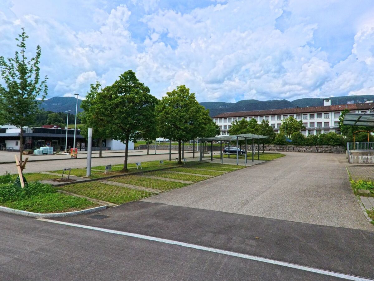 In Langendorf (4513, Kanton Solothurn) liegt ein weitläufiger Parkplatz mit überdachten Fahrradstellplätzen vor einem grossen Gebäude mit Blick auf die Jurakette.