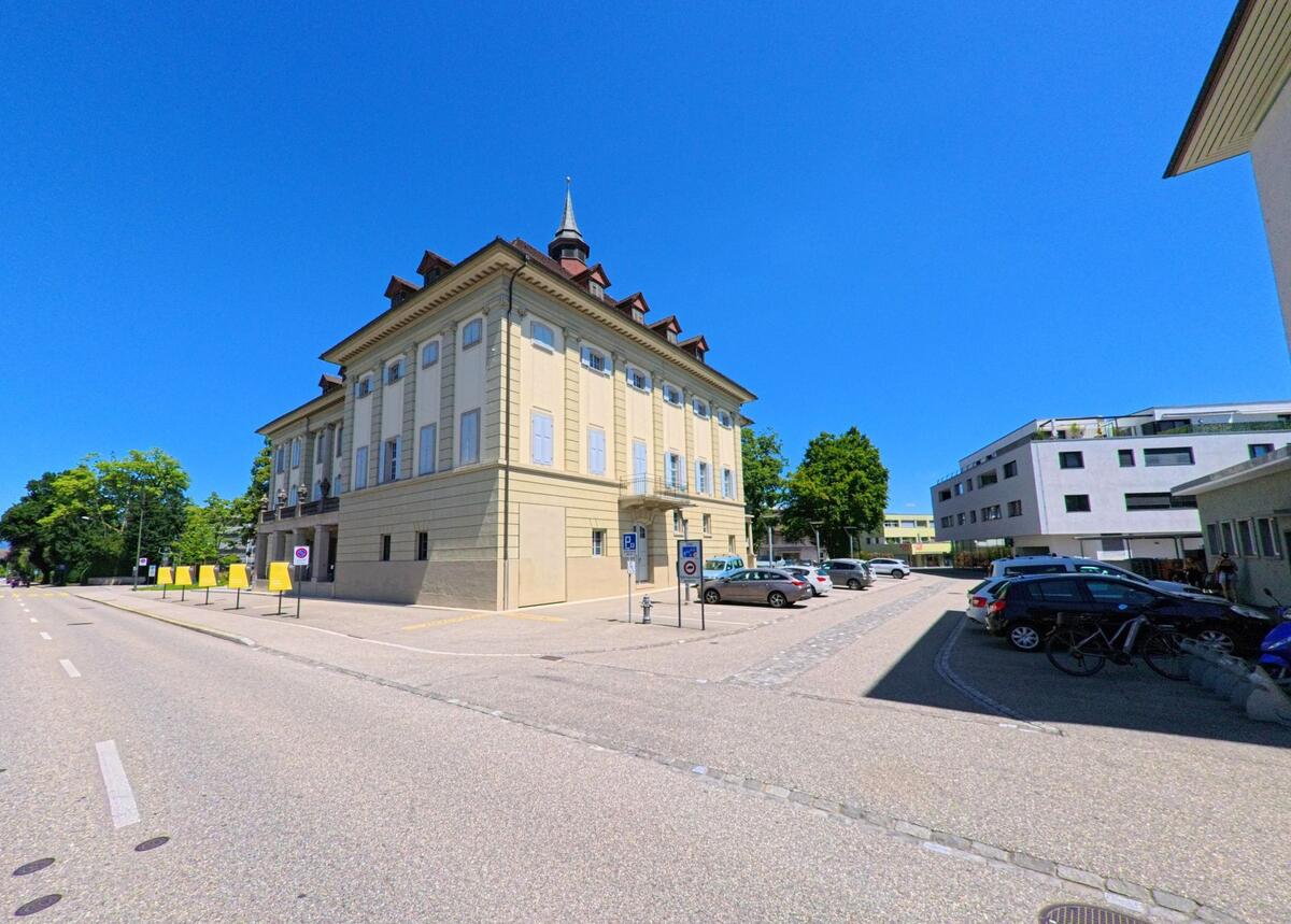 Ein stattliches Gebaeude mit Turmspitze und regelmaessiger Fensterfassade steht in Langenthal (4900, Kanton Bern) an einer breiten Strasse mit angrenzendem Parkplatz.