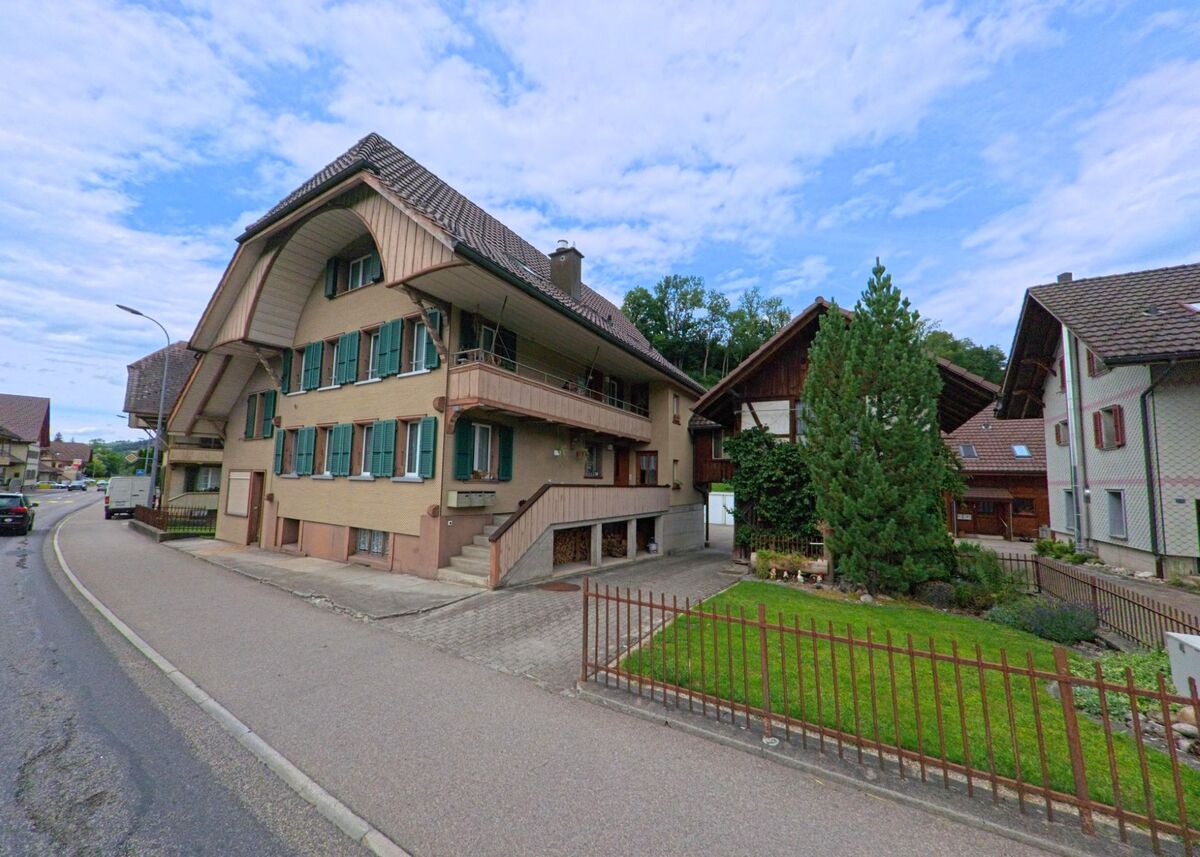 Ein stattliches Emmentaler Wohnhaus mit gruener Fensterlaeden und geschwungenem Dach steht in Langnau im Emmental (3552, Kanton Bern) an einer leicht gekruemmten Strasse mit angrenzendem Garten.