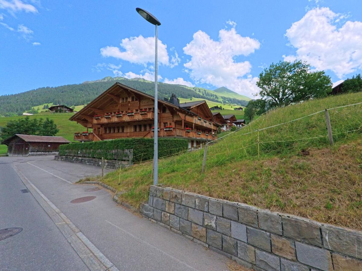 In Lauenen (3782, Kanton Bern) erhebt sich ein grosses Chalet mit Blumenbalkonen vor einem sanften Hang, eingerahmt von Strassenlaterne, Steinmauer und grüner Hügellandschaft.