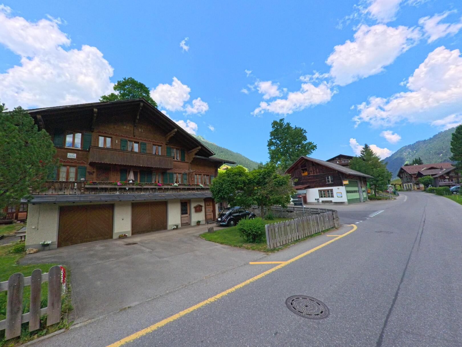 In Lauenen (3782, Kanton Bern) säumen traditionelle Chalets mit Holzbalkonen und Fensterläden eine kurvige Dorfstrasse, eingebettet in eine grüne Berglandschaft.